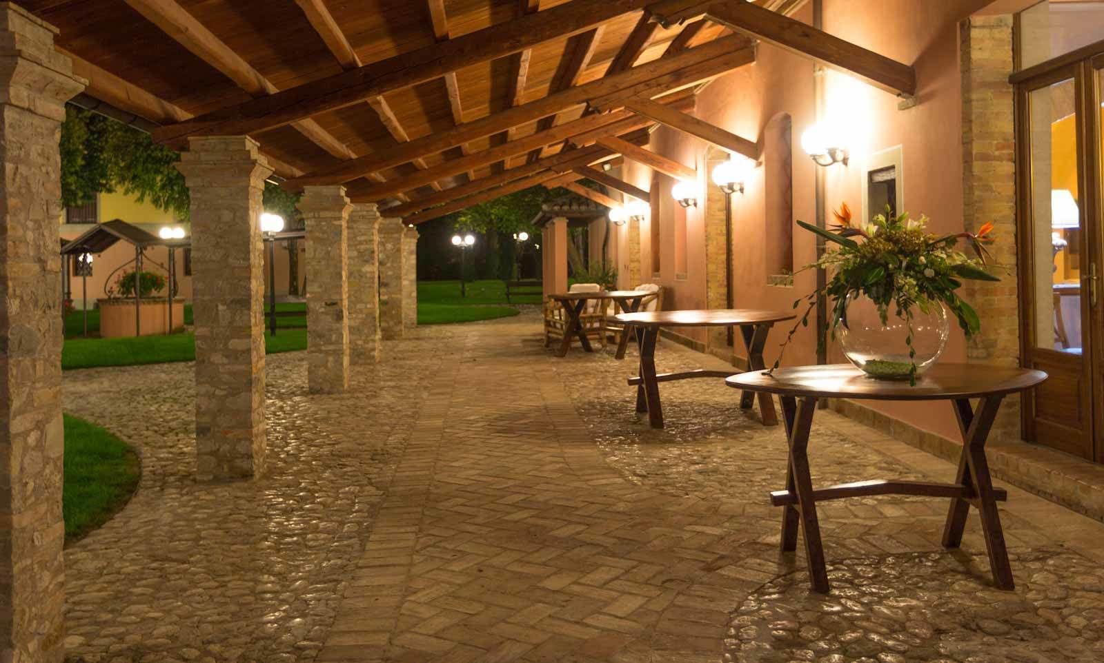 Un patio extérieur couvert, doté d’un sol en pierre, de tables et de chaises en bois, et d’une plante en pot posée sur l’une des tables.