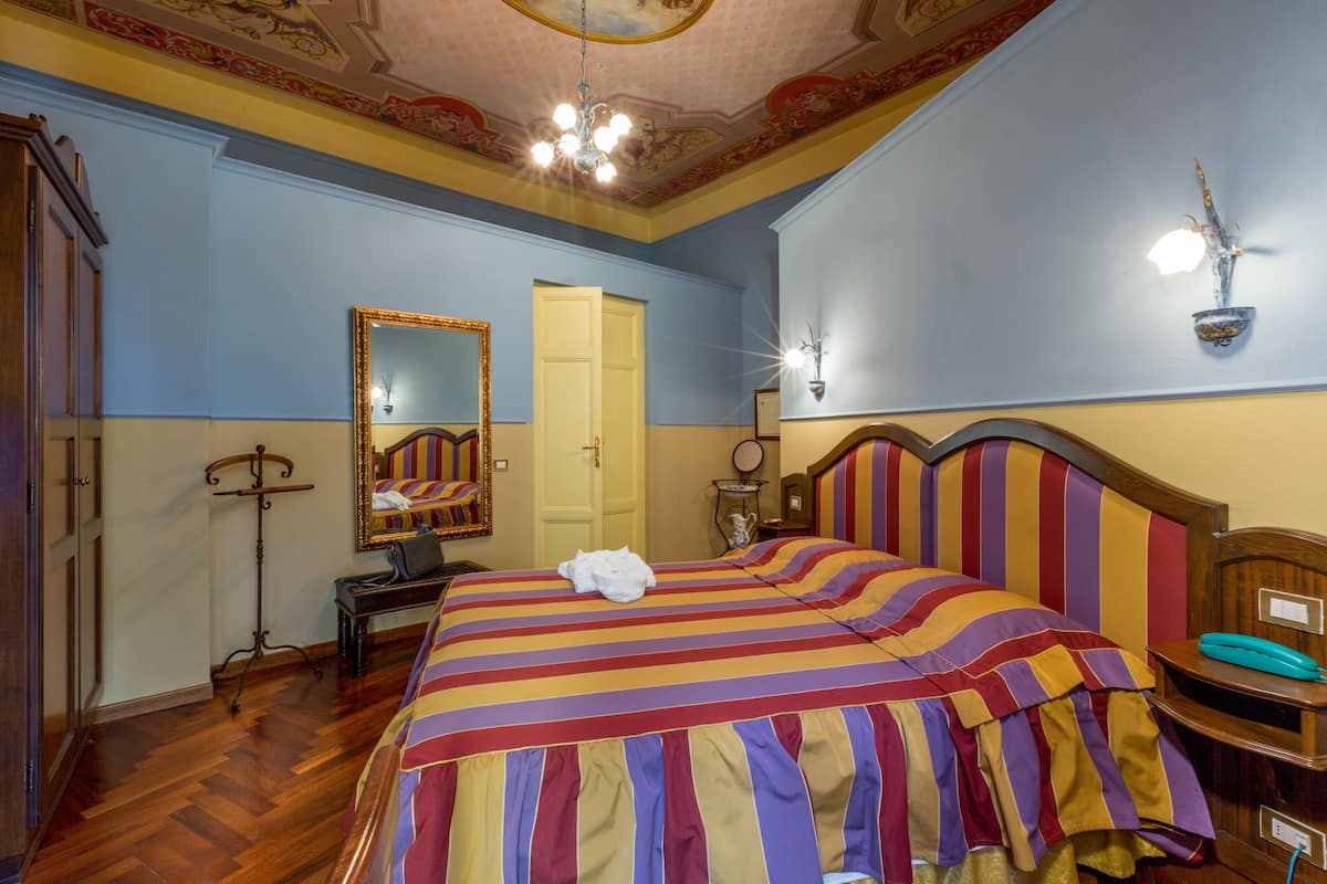 A hotel room with a large bed, a wooden headboard, a bedside table with a telephone, a mirror, and a door leading to another room.
