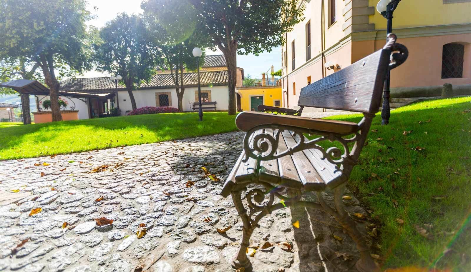 Un camino adoquinado que lleva a un banco con intrincados detalles de metal, rodeado de follaje y edificios de arquitectura tradicional.