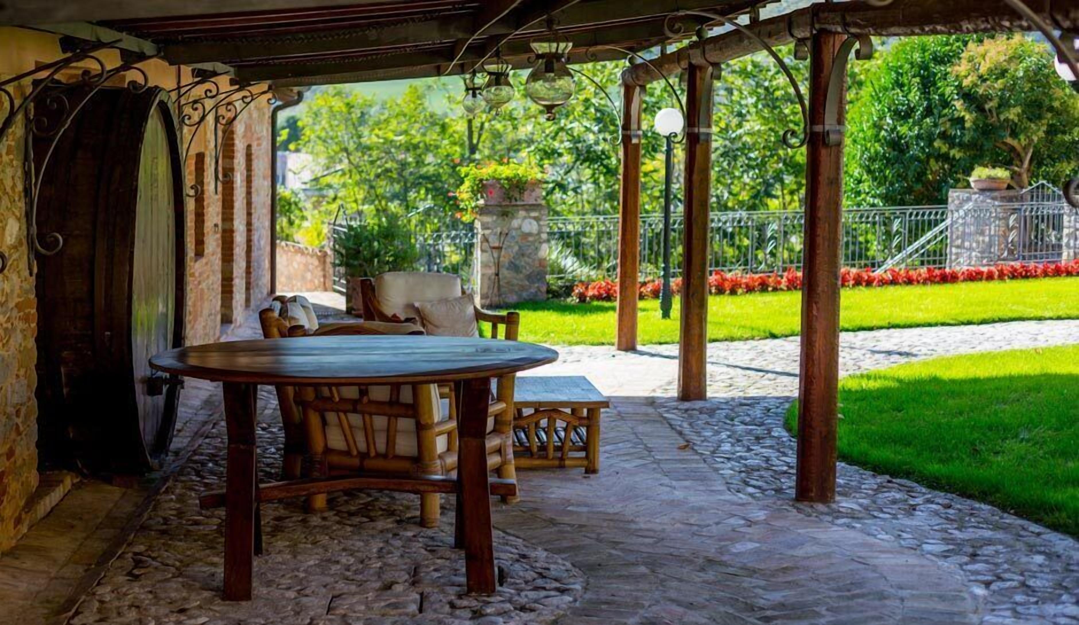 Patio esterno con pavimento in pietra, arredi in legno e vista su un giardino con muro in pietra e fiori rossi.