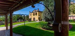 A building with a central arched entrance, surrounded by a well-maintained garden with a gravel path leading to it. - Corte dei Tini (Teramo)