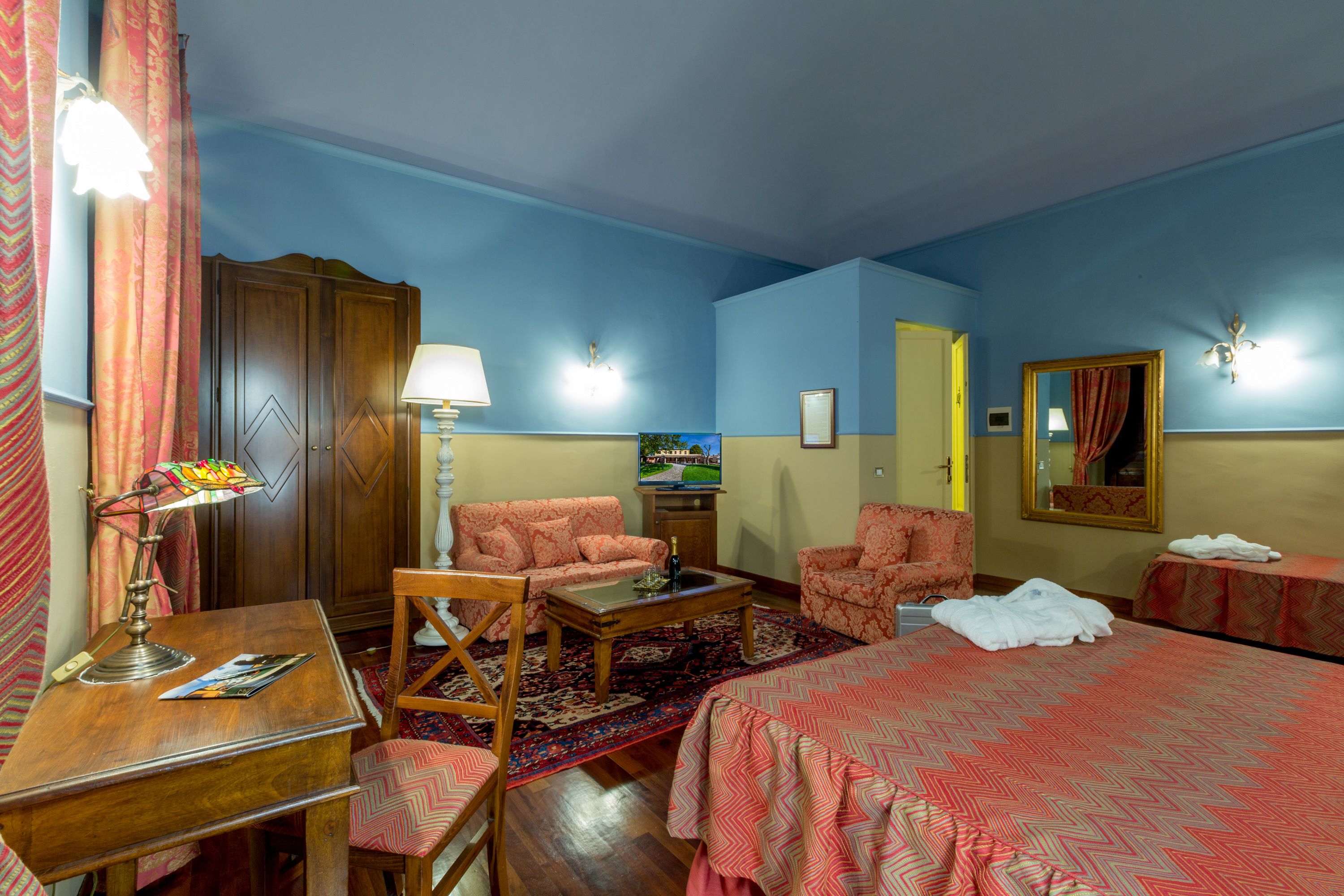 Una camera d'albergo con un letto, due poltrone, una scrivania in legno, una lampada, una televisione e un tappeto decorato.