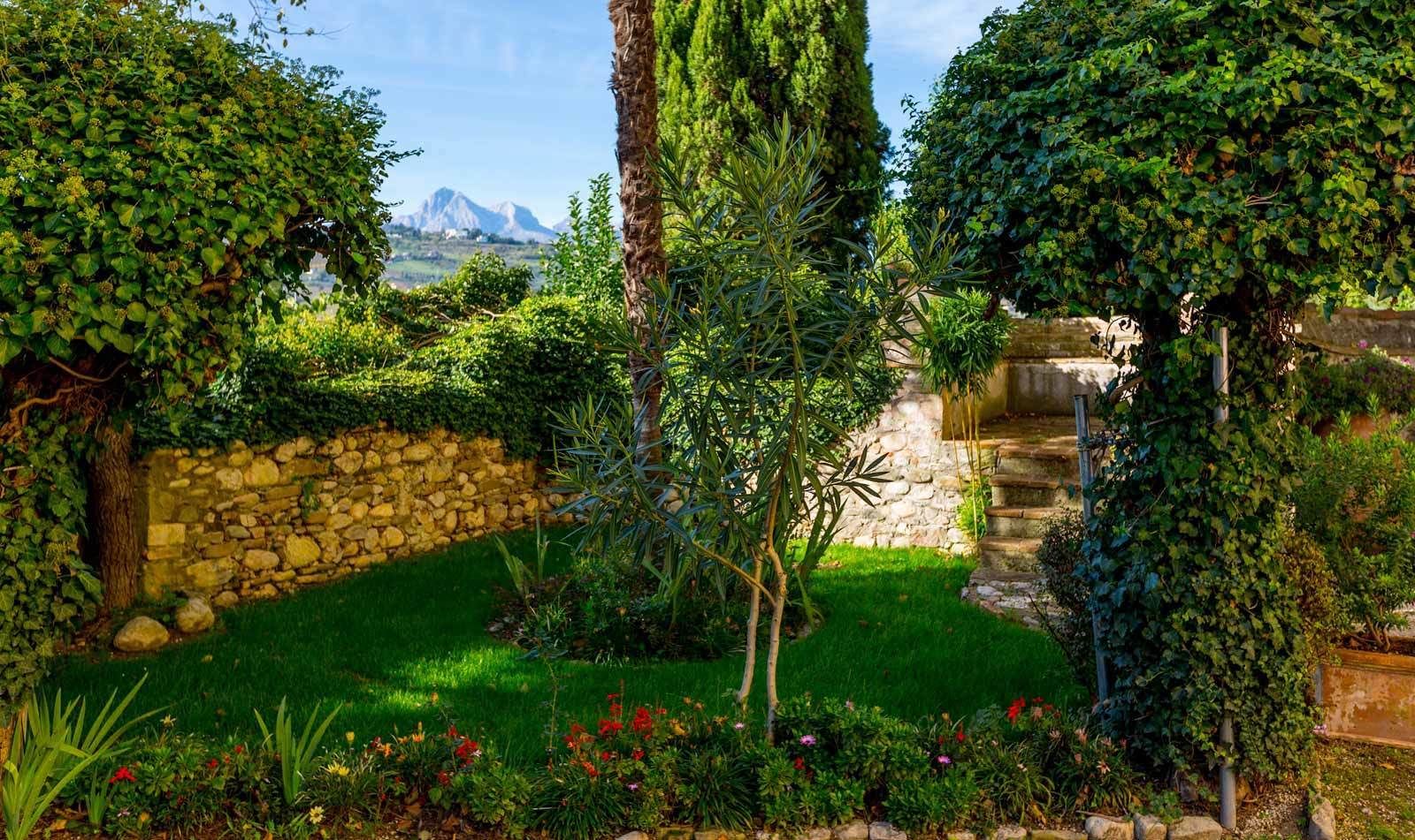 Un jardin luxuriant, avec un mur de pierre, un palmier et un escalier menant à une construction en pierre.