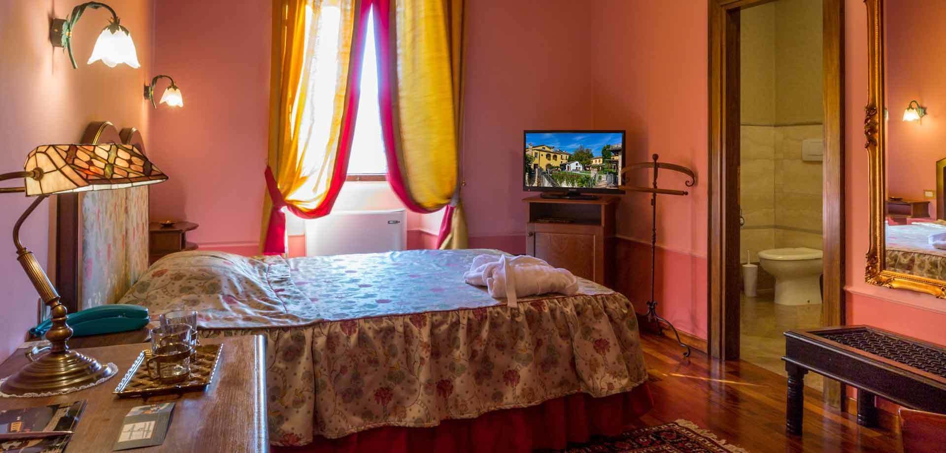 Une chambre à coucher avec un lit, un bureau avec une lampe, une télévision et une salle de bain visible à travers une porte ouverte.
