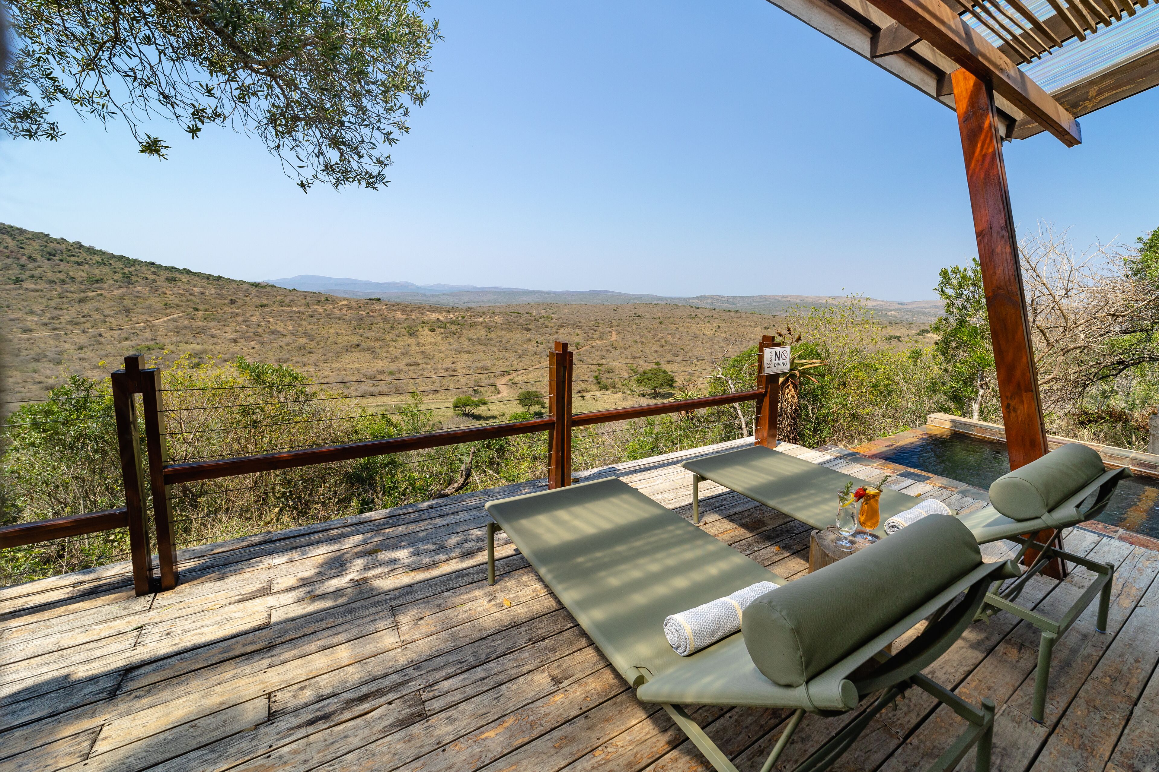 A wooden deck with two lounge chairs and a small table, overlooking a dry landscape with sparse vegetation and distant hills.