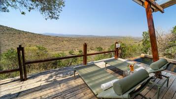 A wooden deck with two lounge chairs and a small table, overlooking a dry landscape with sparse vegetation and distant hills.