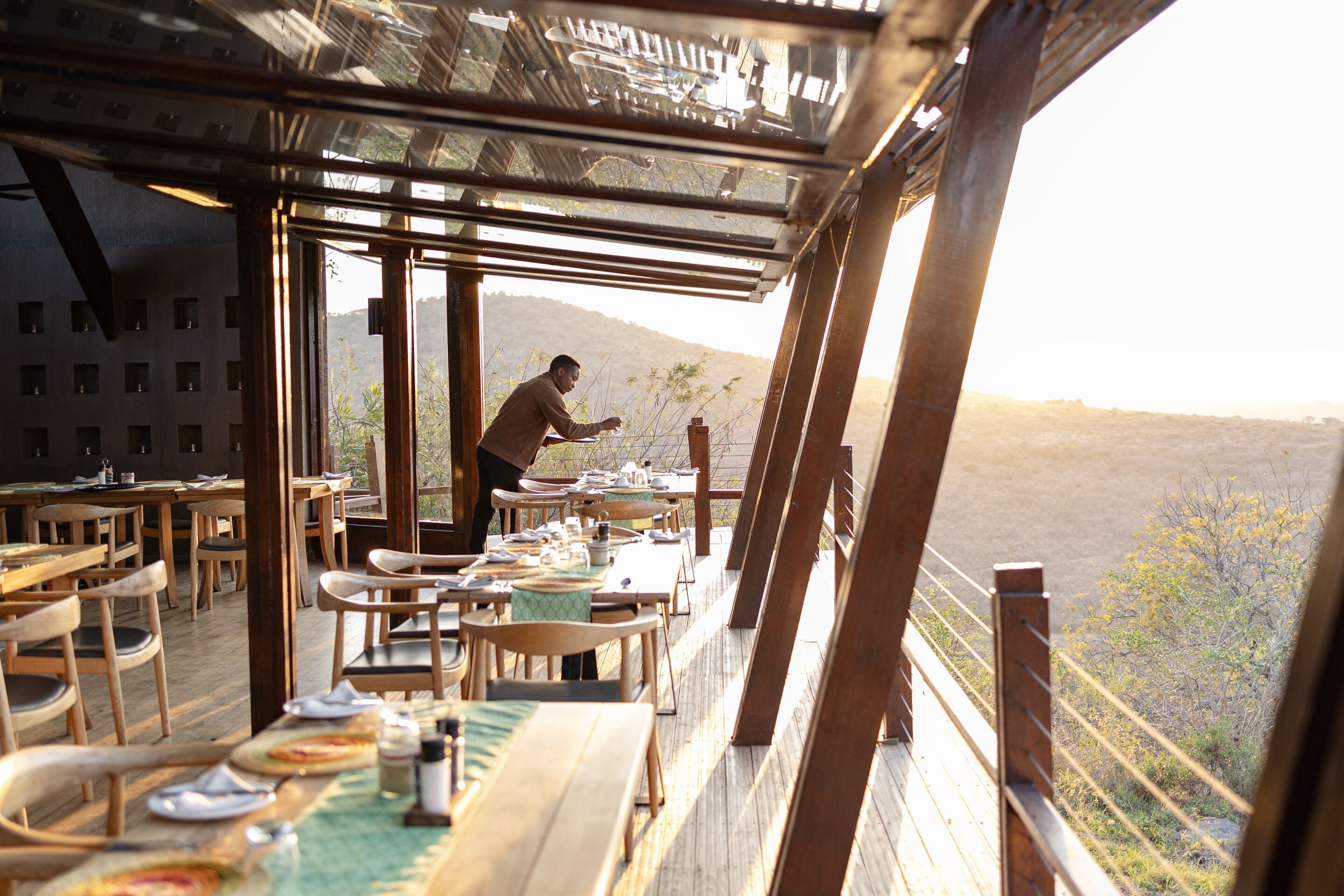 An outdoor dining area with wooden tables and chairs, a person serving food, and a view of a mountainous landscape.
