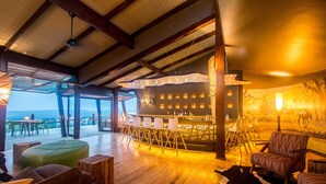 Ein geräumiger Raum mit Holzboden, einer Bar mit Hockern, einem Kuhfellteppich und einem großen Fenster mit Meerblick.