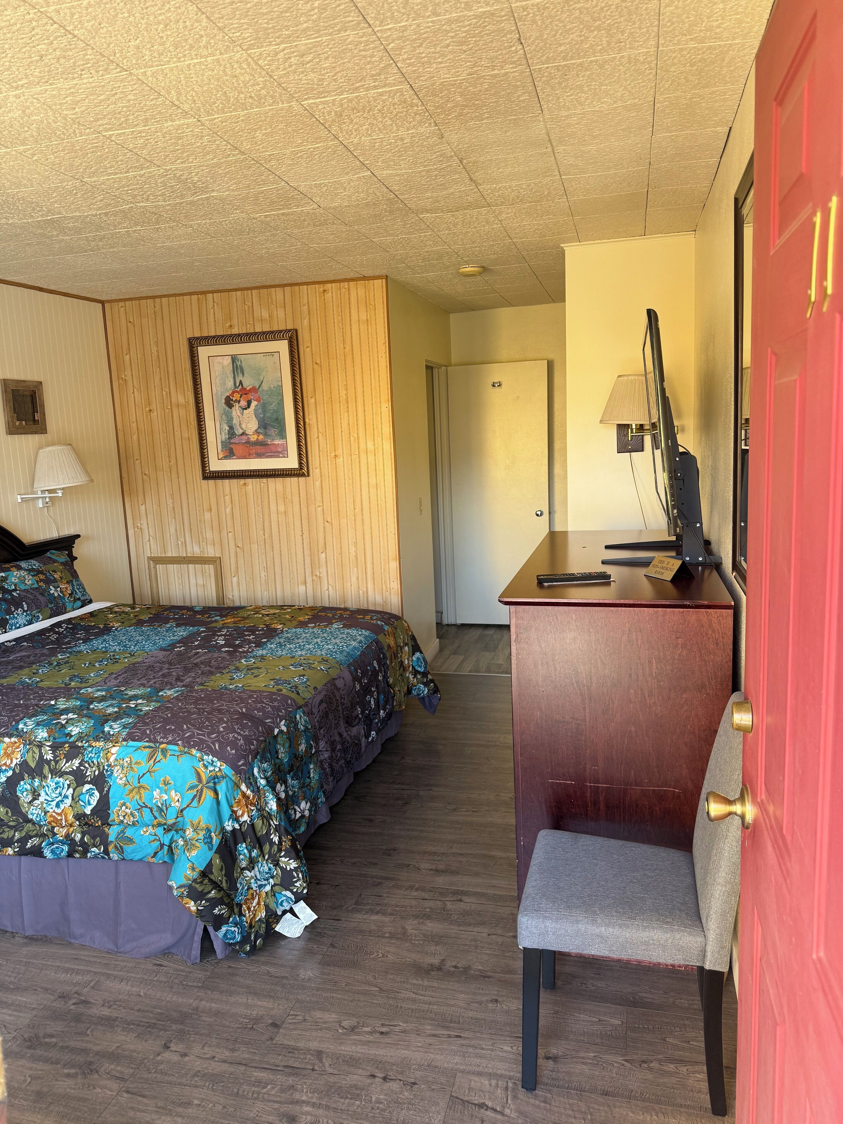 A hotel room with a bed, a desk with a computer, a chair, a lamp, and a door.
