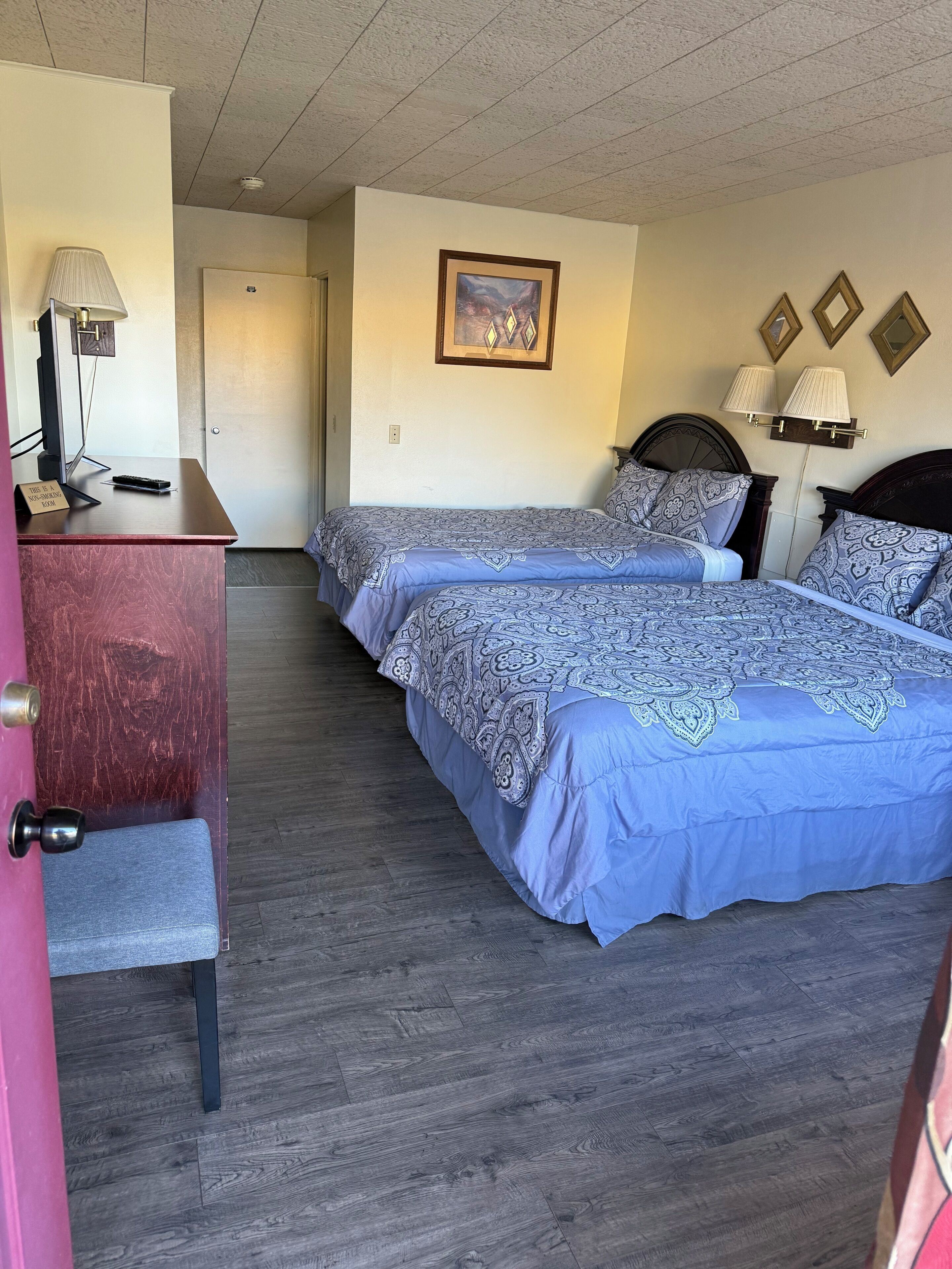 A hotel room with two beds, a desk, a lamp, and a painting on the wall.