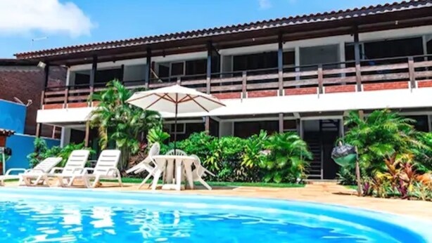 A swimming pool with lounge chairs and umbrellas, surrounded by lush greenery and a building with a tiled roof. - Pousada do Frade (Ipojuca)
