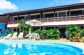 A swimming pool with lounge chairs and umbrellas, surrounded by lush greenery and a building with a tiled roof. - Pousada do Frade (Ipojuca)