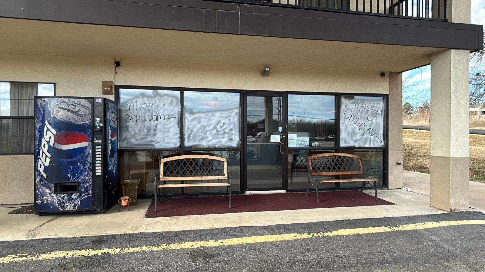 A building with a covered seating area, two wooden benches, a Pepsi vending machine, and a glass door with a 'Merry Christmas' sign.