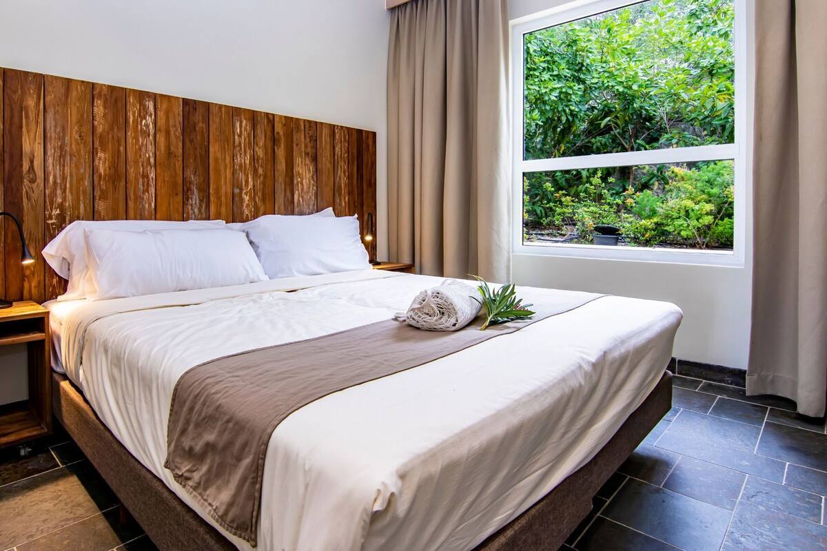 Una cama bien hecha con sábanas blancas y un plaid marrón, una mesita de noche con una lámpara, y una ventana con vistas a la vegetación.