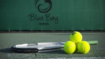 Una raqueta de tenis y tres pelotas de tenis en una cancha con un muro verde al fondo.