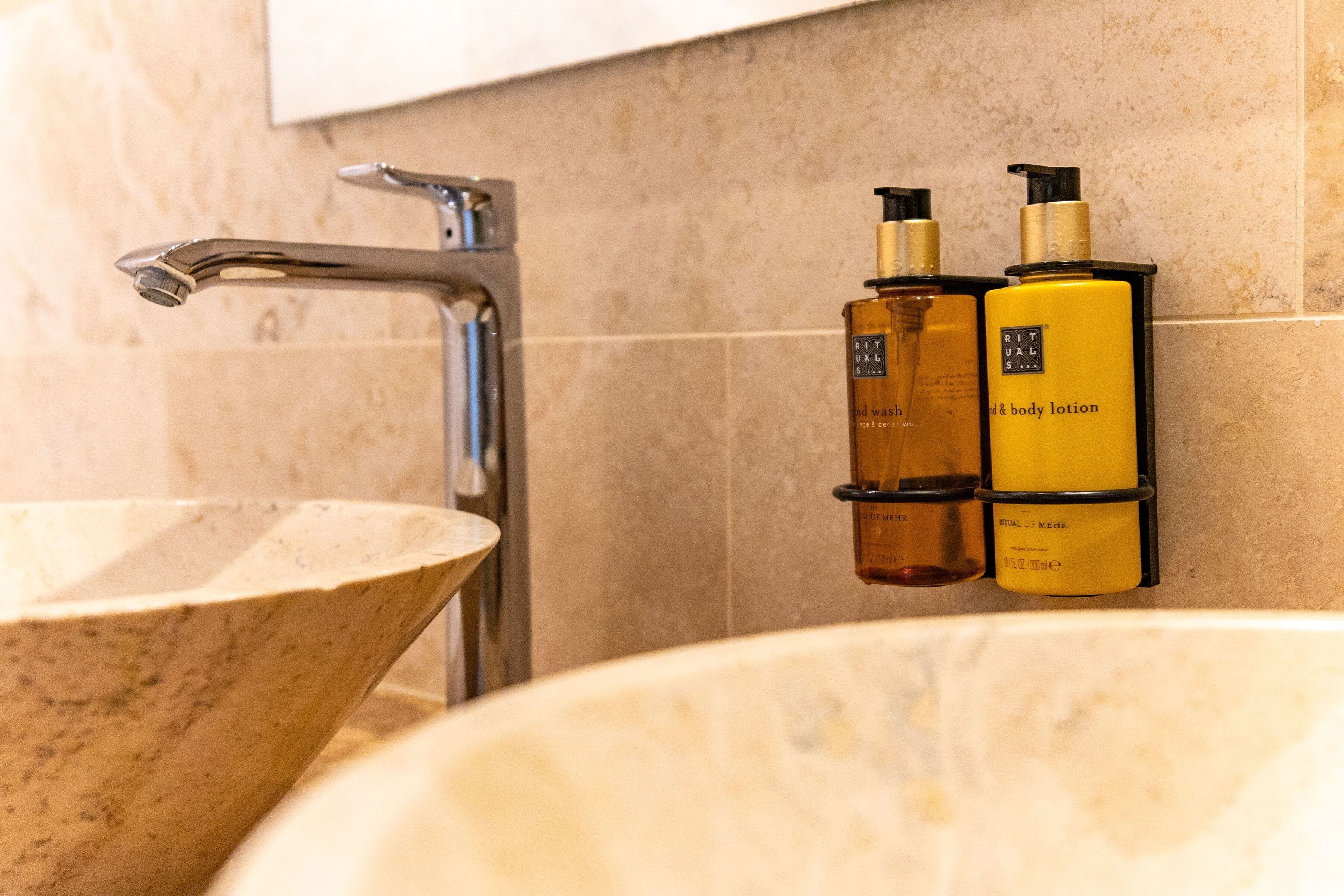 Een badkamer met een marmeren wastafel, een moderne kraan en twee flesjes bodylotion op een wandhouder.