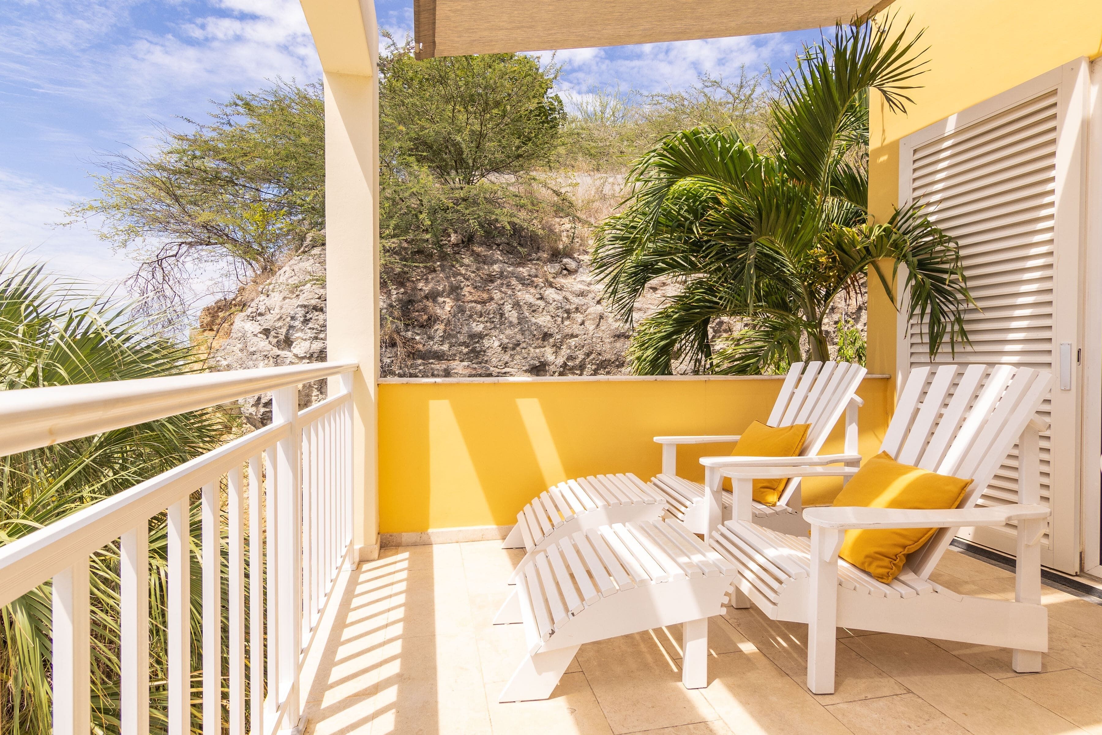 A balcony with white lounge chairs and yellow cushions, overlooking a rocky hillside and palm trees.