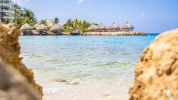A beach with clear turquoise water, thatched-roof structures, and a building with balconies. - Bayside Boutique Hotel - Blue Bay Golf & Beach Resort (Sint Michiel)