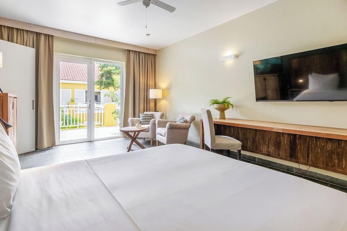 A hotel room with a bed, a desk, a chair, a TV mounted on the wall, and a balcony with curtains.