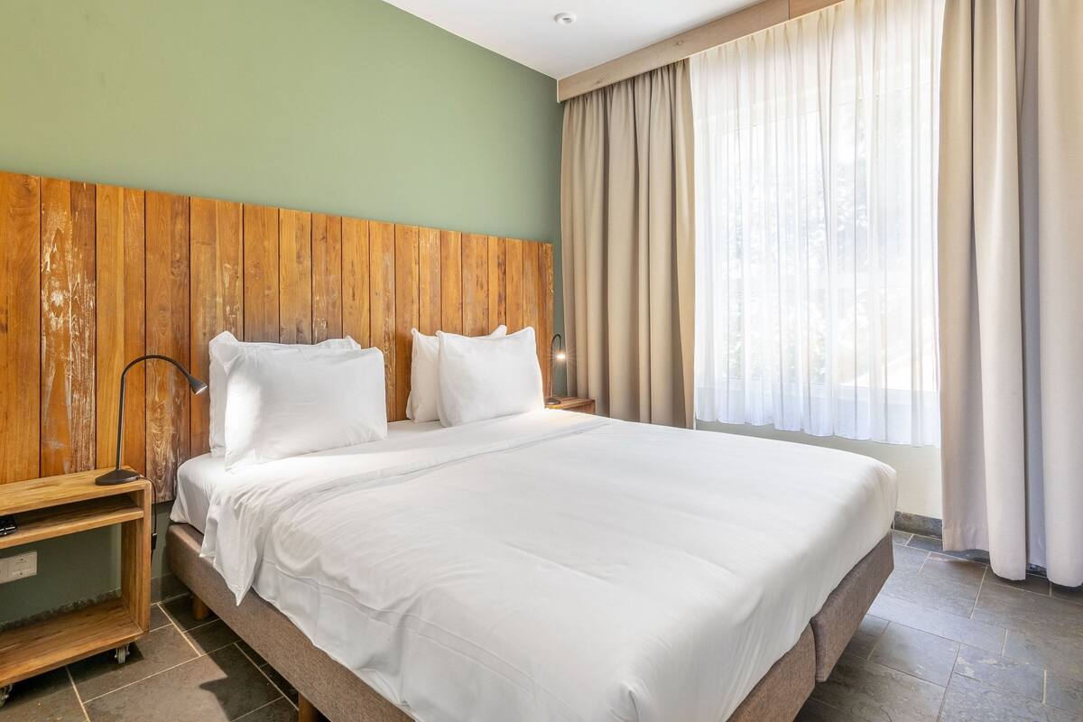 Una cama bien tendida con ropa de cama blanca en una habitación con cabecera de madera, una mesita de noche y una ventana con cortinas.