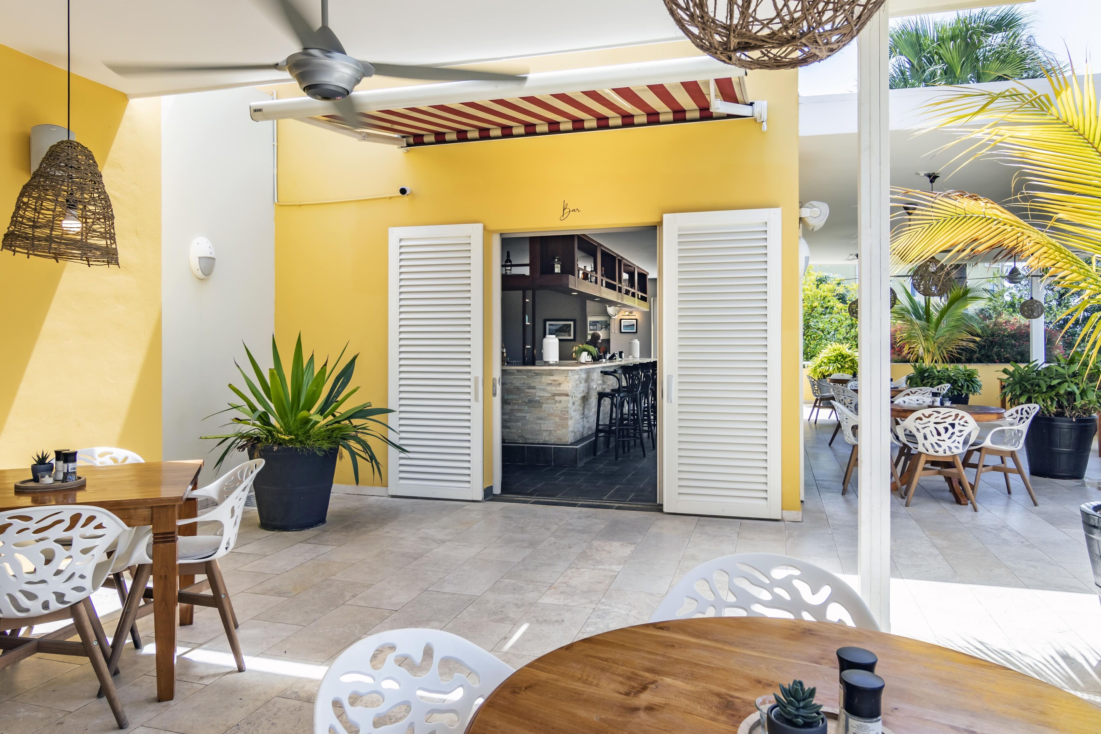A bright yellow room with white shutters, a ceiling fan, and a view of a bar area with outdoor seating.