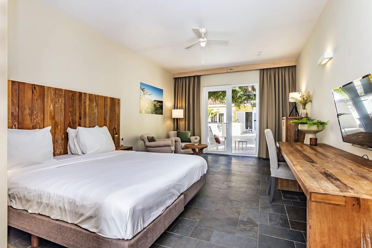 A hotel room with a large bed, a desk, a chair, a ceiling fan, and a view through the glass door.