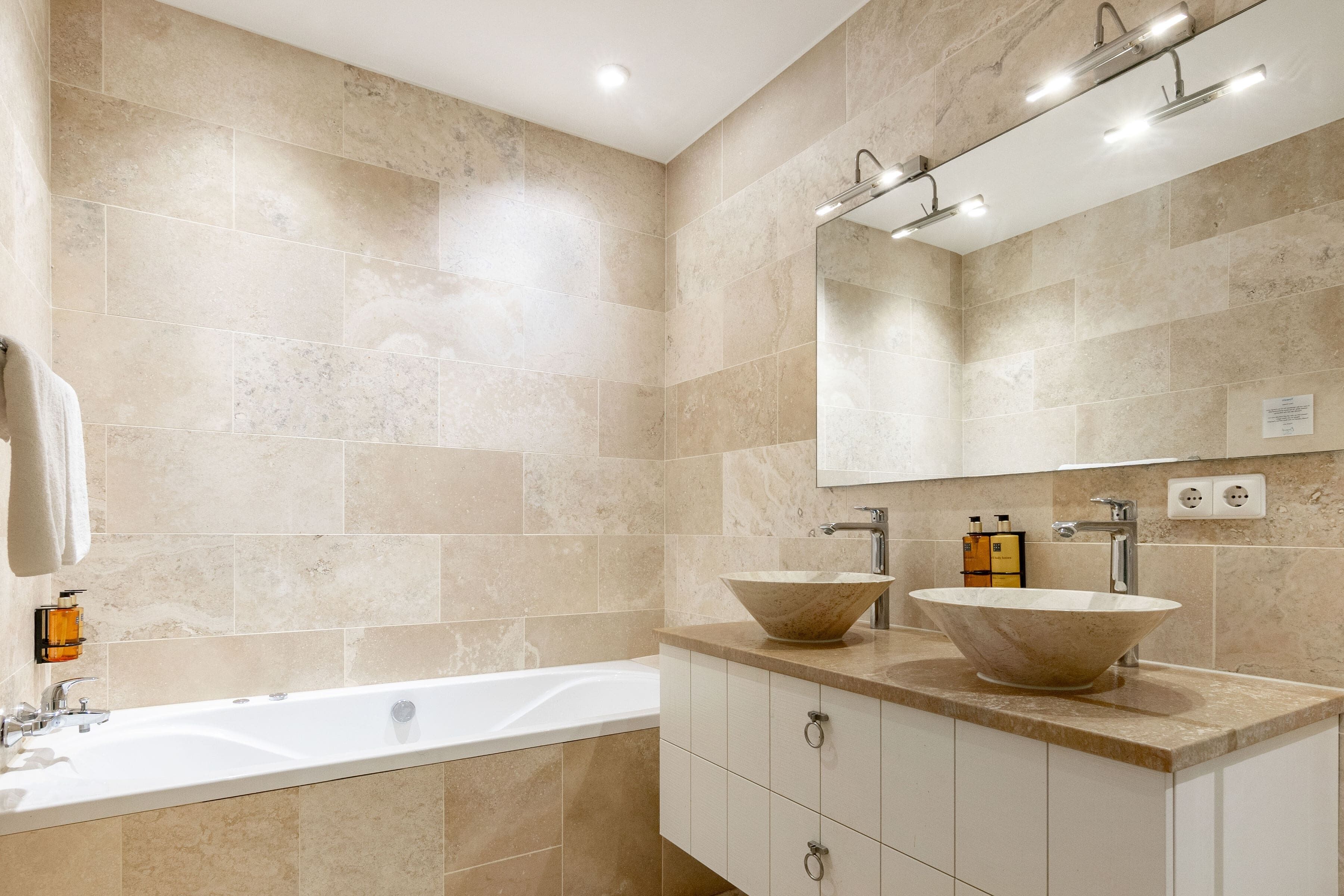 A modern bathroom with beige tiles, a large bathtub, two vessel sinks, and a mirror above the sink.