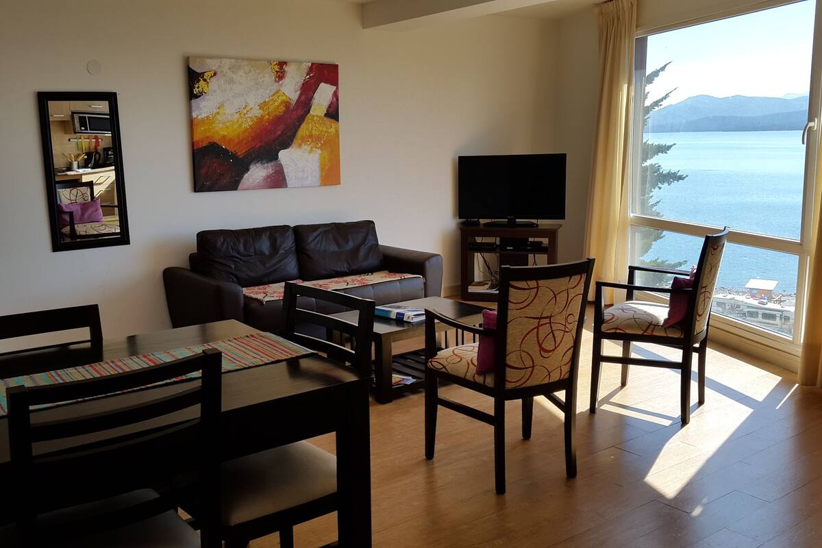 Un salon comprenant un canapé, un téléviseur, une table à manger avec des chaises et une grande fenêtre donnant sur la mer.