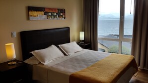 Een hotelkamer met een bed, een nachtkastje met een lamp, een schilderij aan de muur en uitzicht op de zee.