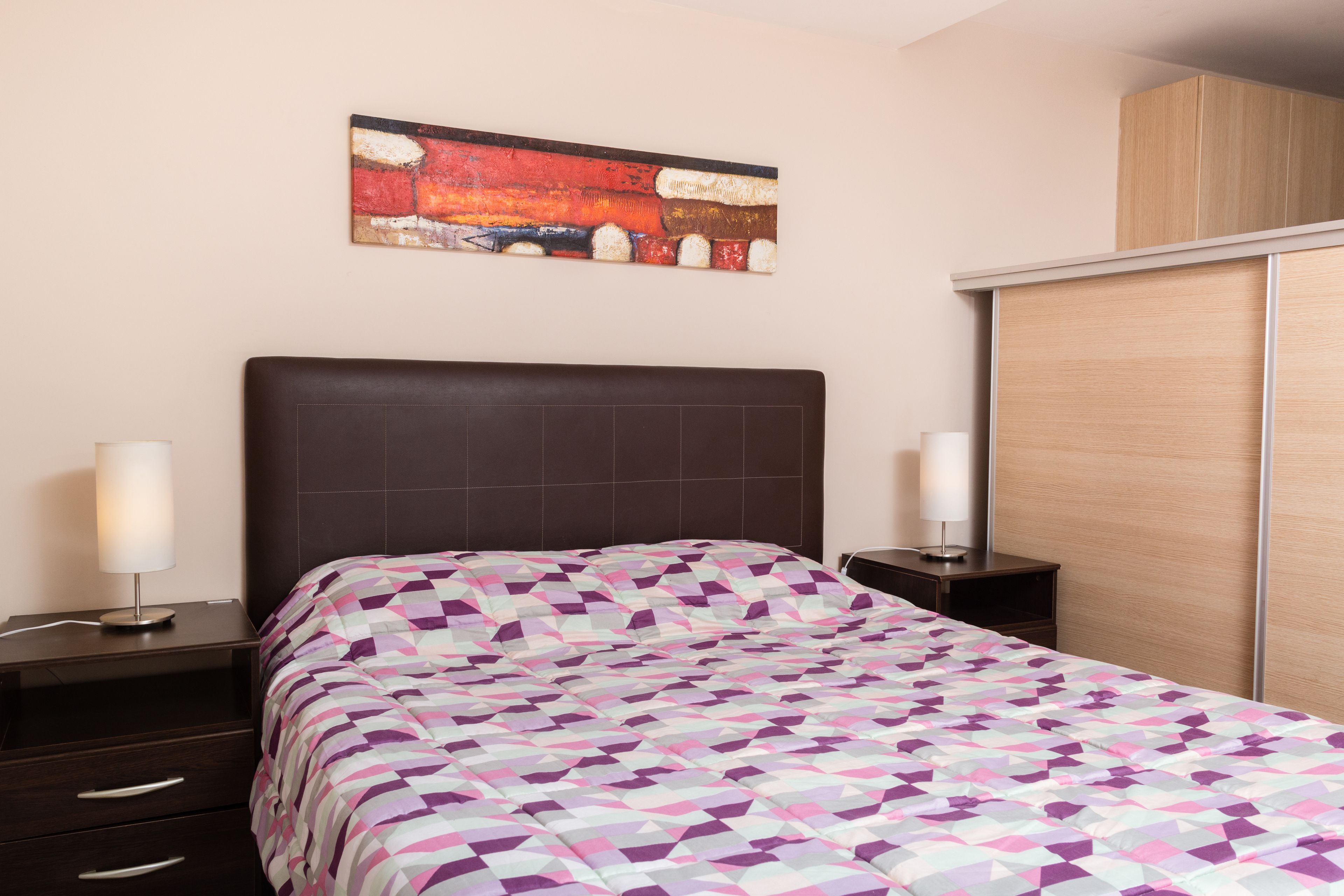 Un dormitorio con cama, mesitas de noche, un armario de madera y un cuadro en la pared.