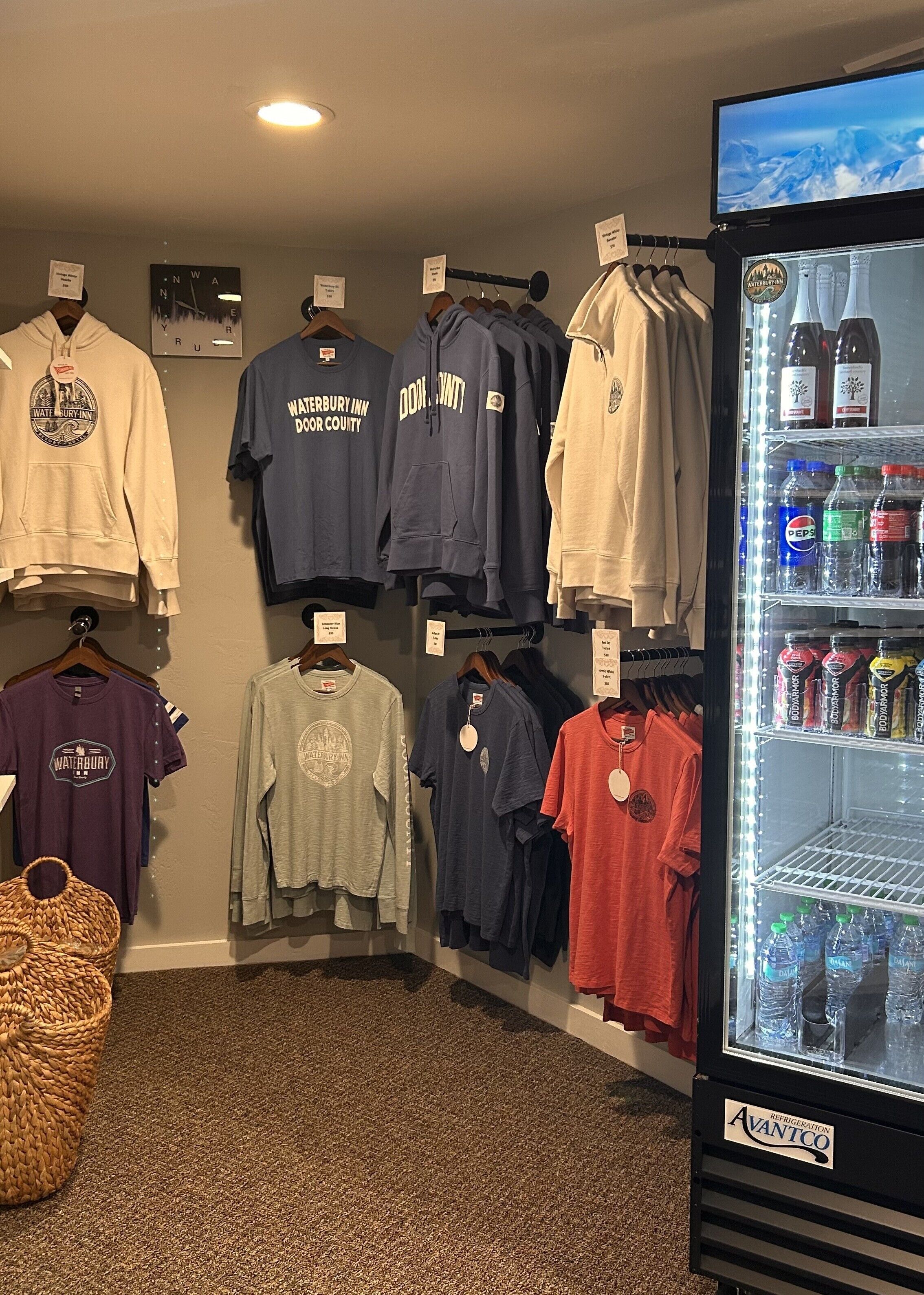 A retail display with various Waterbury Inn DOOR COUNTY branded clothing and a refrigerator stocked with beverages.