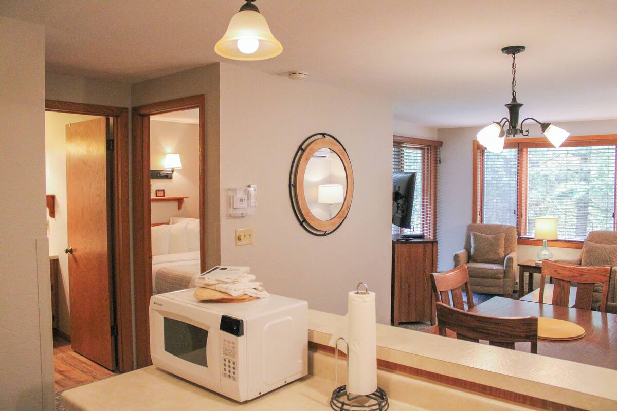 A hotel room with a kitchenette, a dining area with a table and chairs, a mirror, and a ceiling light fixture.