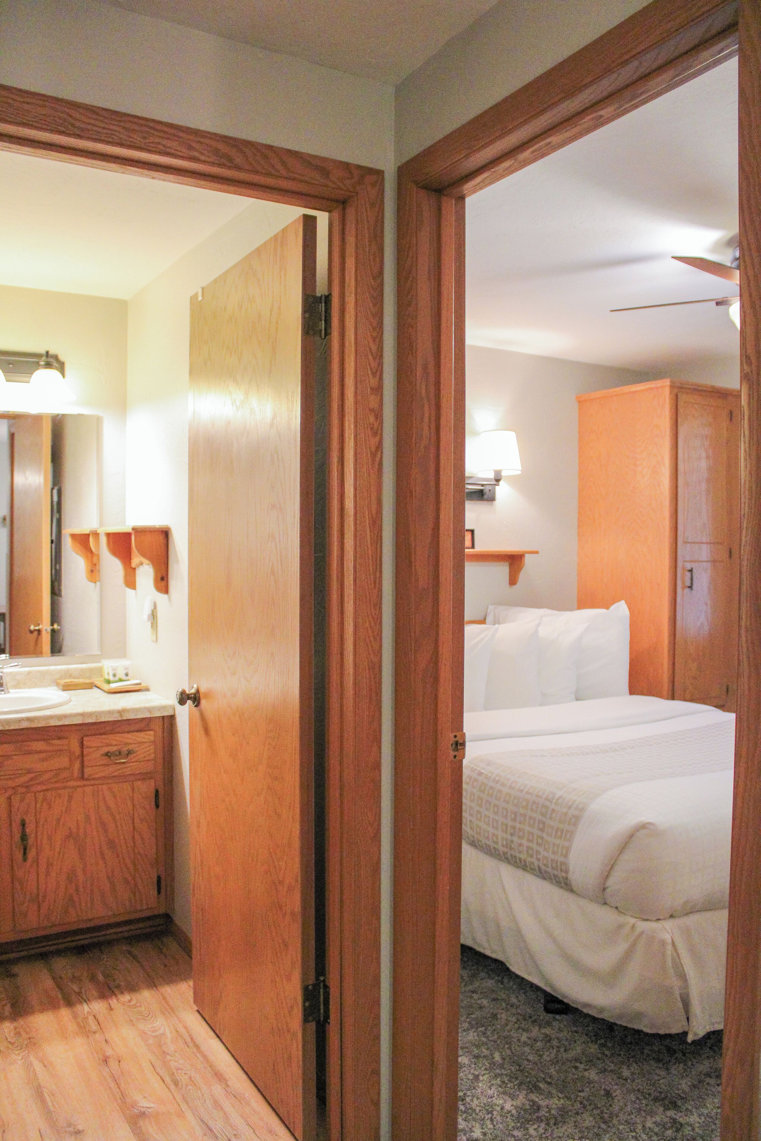 A hotel room with a wooden door, a bed with white bedding, a bedside table, a mirror, and a wooden wardrobe.