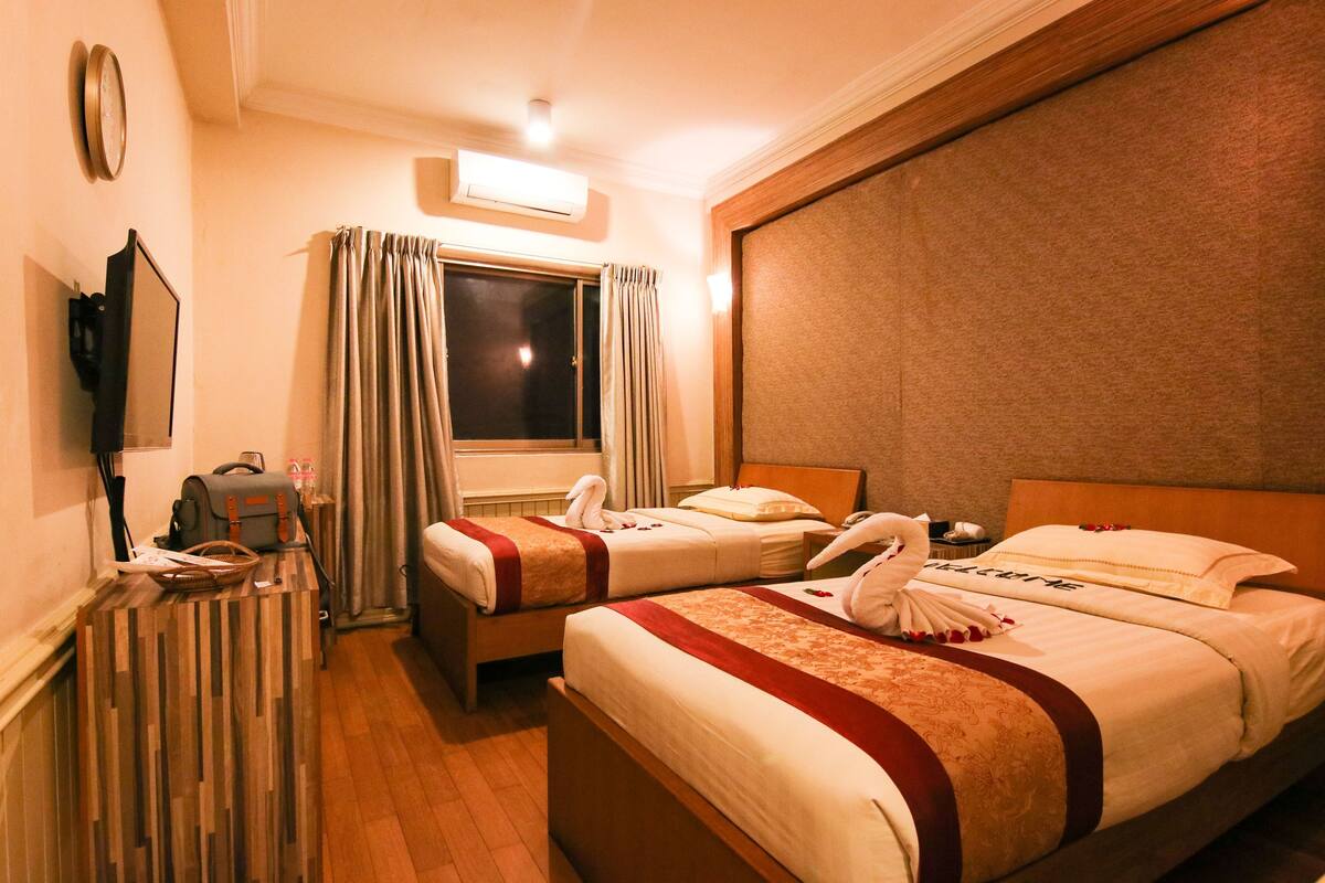 Ein Hotelzimmer mit zwei Betten, einem Schreibtisch mit Lampe, einem Fenster mit Vorhängen und einer Wanduhr.