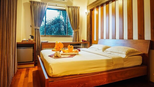 Uma cama bem arrumada com travesseiros e um cobertor, uma mesa de cabeceira de madeira com uma luminária, uma janela com cortinas e vista para árvores lá fora.