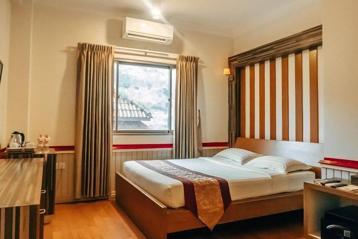 Ein Hotelzimmer mit einem Bett, einem Schreibtisch, einem Sessel, einem Fenster mit Vorhängen, einer Klimaanlage und einem Fernseher.