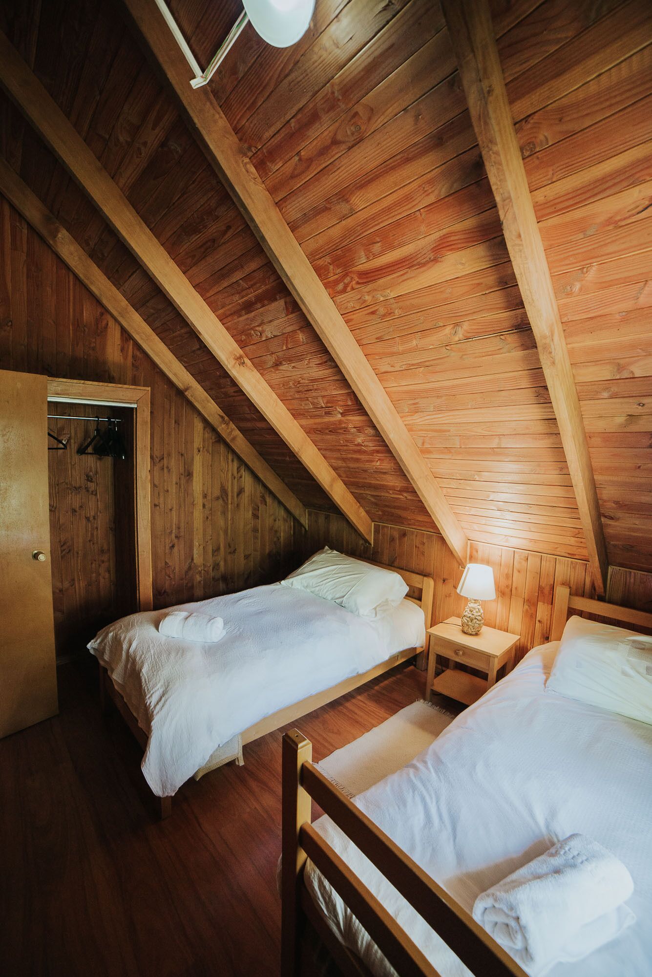 Ein gemütliches Schlafzimmer im Dachgeschoss mit Holzwänden und schrägen Decken, mit zwei Betten und einem Nachttisch mit Lampe.