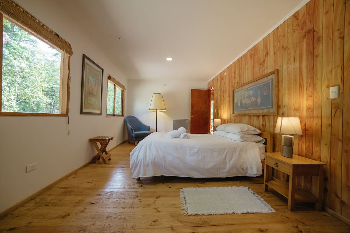 A bedroom with a wooden floor, a bed with white linens, a chair, a bedside table with a lamp, and a painting on the wall.