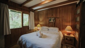 A wooden-paneled bedroom with a bed, two bedside lamps, a window with curtains, and a wall hanging. - Cabañas Metreñehue (Pucón)