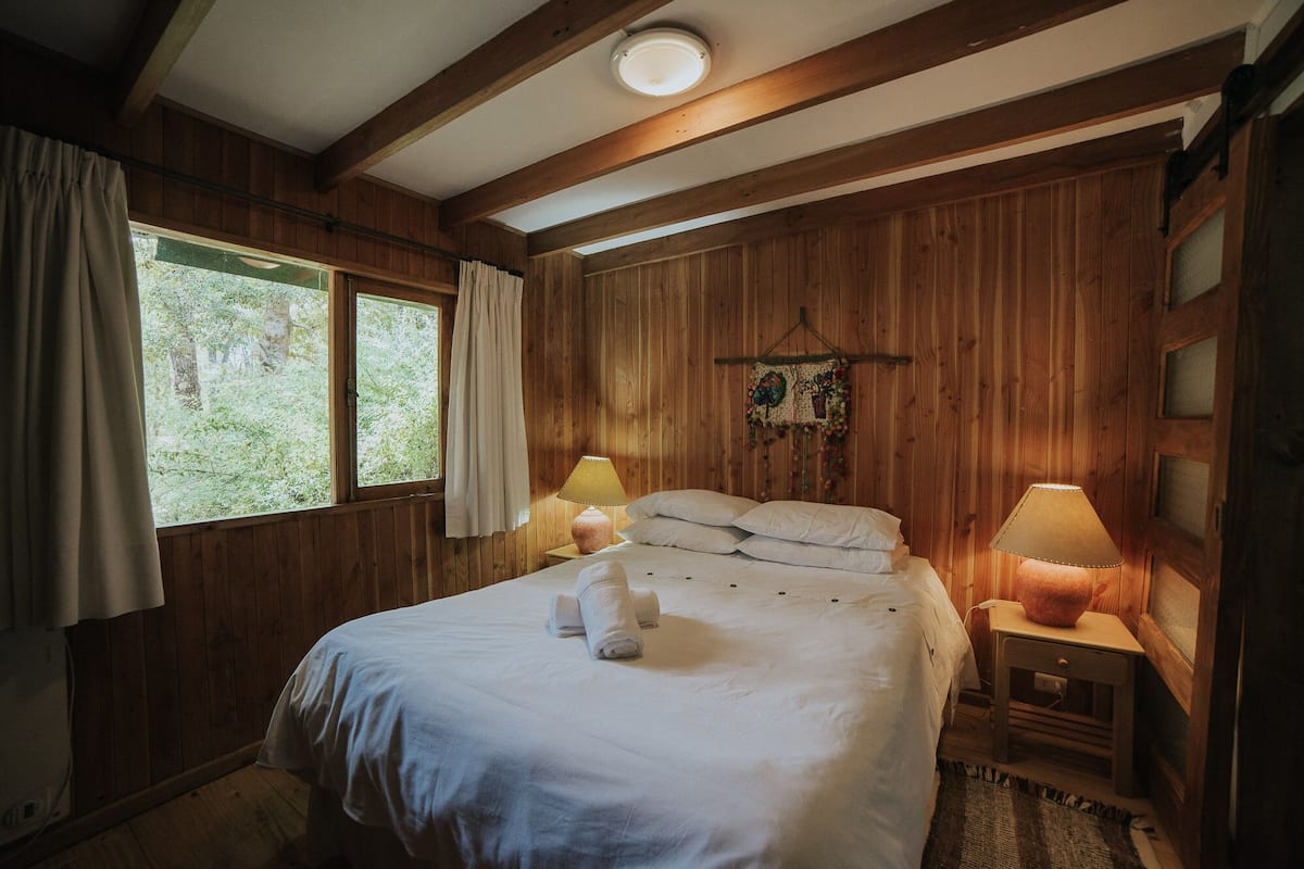 A wooden-paneled bedroom with a bed, two bedside lamps, a window with curtains, and a wall hanging.
