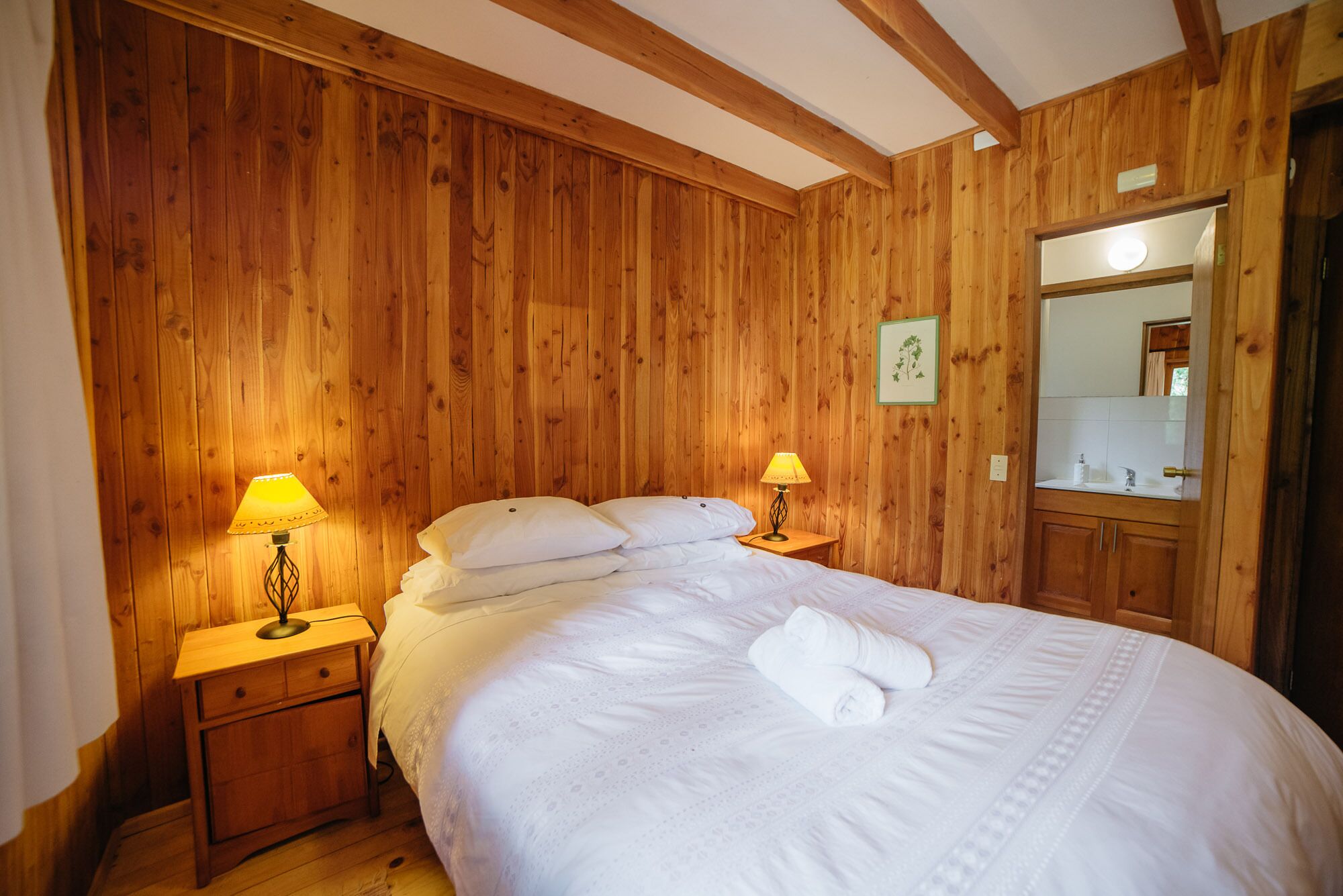 A wooden-paneled bedroom with a bed, two bedside lamps, a mirror, and a framed picture on the wall.