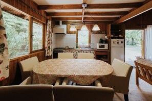 A rustic kitchen with wooden beams, a round table with floral tablecloth, and a view of greenery outside. - Cabañas Metreñehue (Pucón)