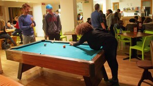 A pool table in a room with green chairs and tables, surrounded by people. - Safestay Pisa Centrale (Pisa)