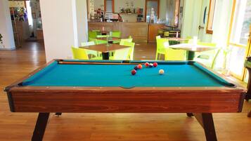 Una mesa de billar con bolas en una sala con suelo de madera, sillas verdes y una zona de cocina al fondo.