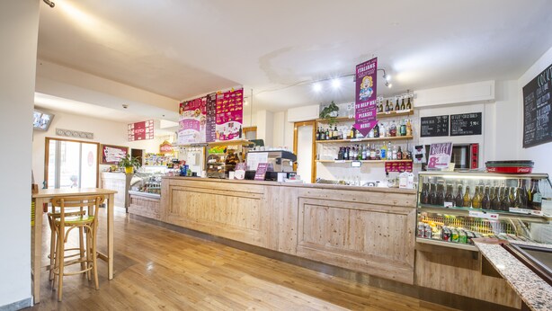 A bar area with wooden furniture, a counter with various bottles, and hanging promotional signs. - Safestay Pisa Centrale (Pisa)