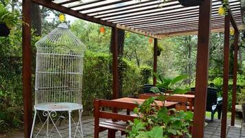 Un área de estar al aire libre con techo, una jaula de pájaros, bancos de madera y plantas en macetas.
