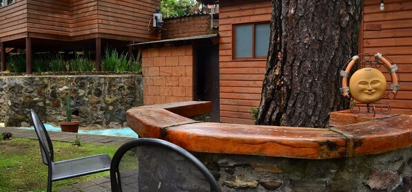 A wooden cabin with a stone wall, a bench, and a tree.