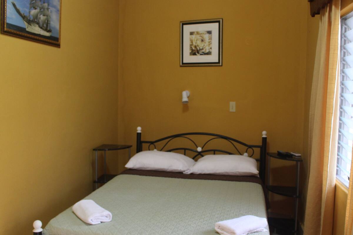 A single bed with a green bedspread and white pillows, a small bedside table, a wall-mounted light, and a framed picture above the bed.