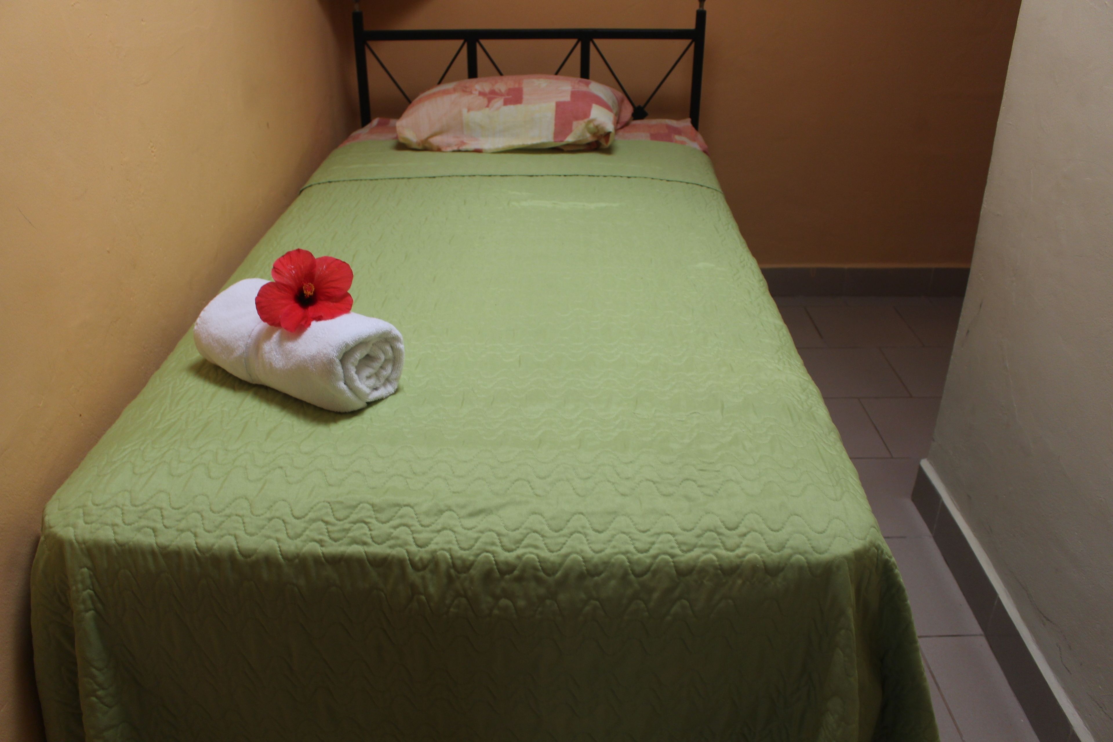Un lit simple avec une couvre-lit verte et une fleur rouge posée sur une serviette blanche.