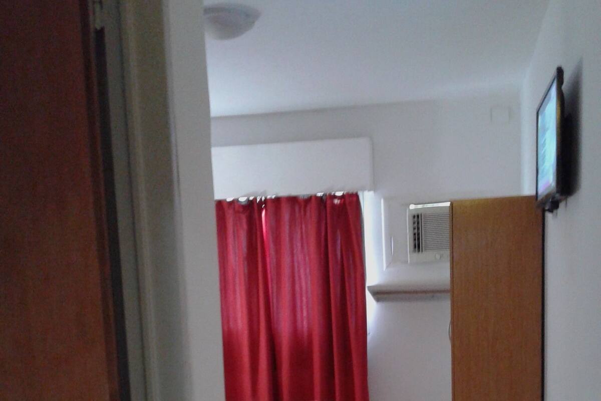 Una habitación pequeña y estrecha con una cama, una cortina roja, un equipo de aire acondicionado blanco y una puerta de madera.
